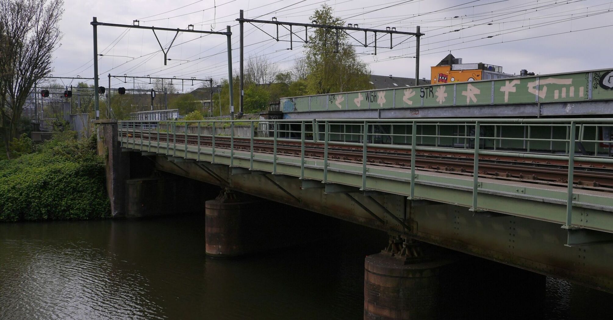 A railway bridge over a river with train tracks and power lines visible, surrounded by greenery