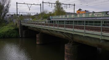 A railway bridge over a river with train tracks and power lines visible, surrounded by greenery