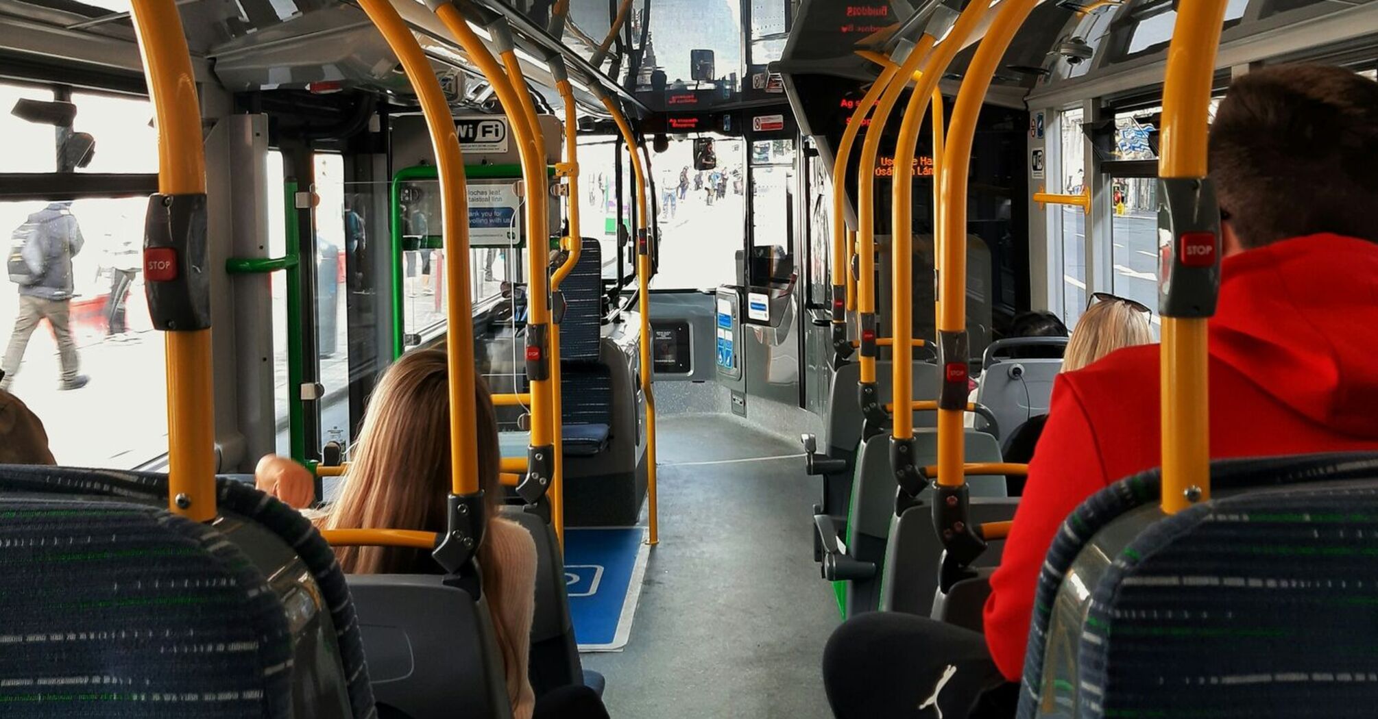 Passengers seated inside a modern public bus, with visible yellow handrails and a clear view of the bus driver's area at the front