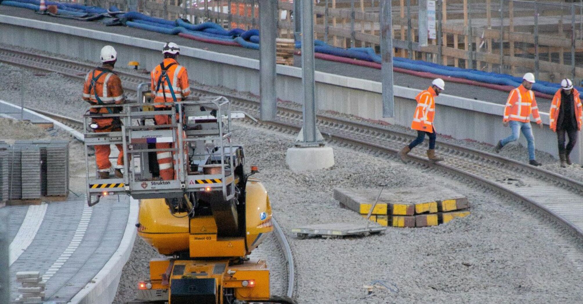 Construction workers in orange vests working on railway track upgrades with machinery on site