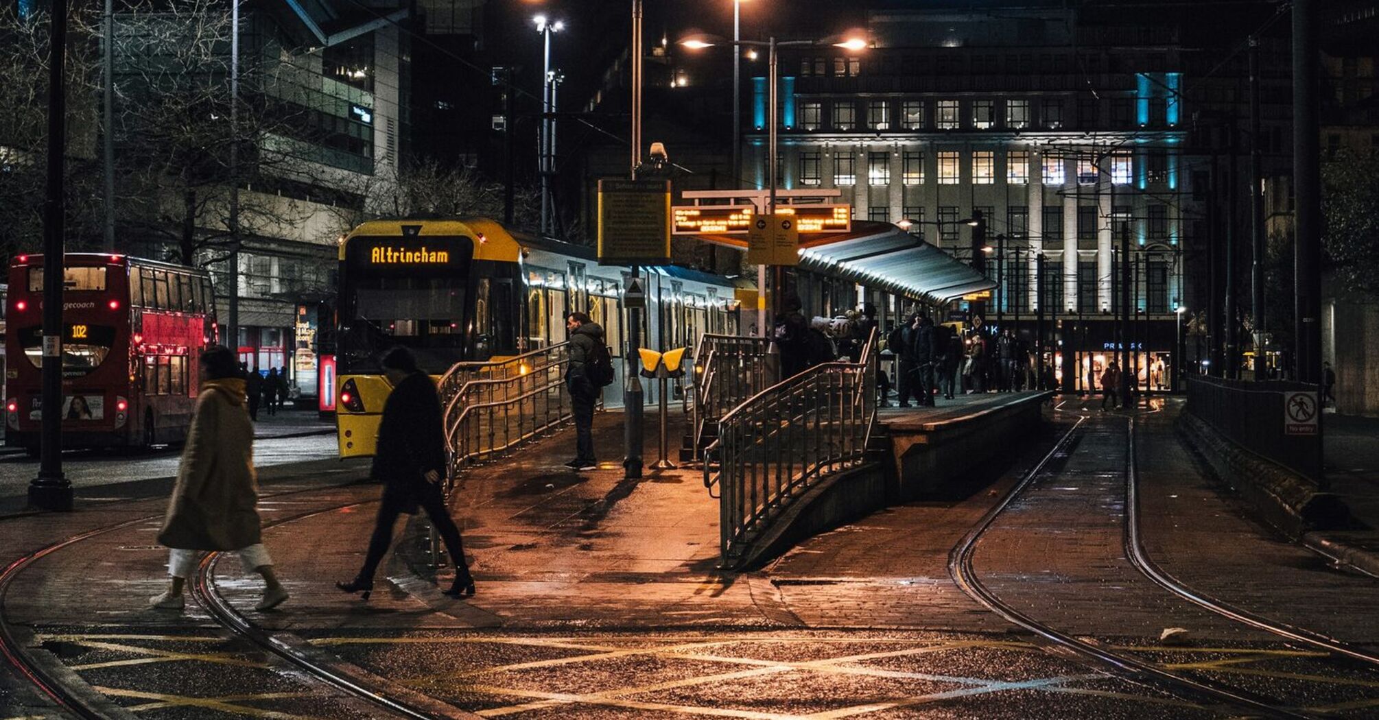 Night scene at a busy tram stop in Manchester with people crossing the street and a bus on the left
