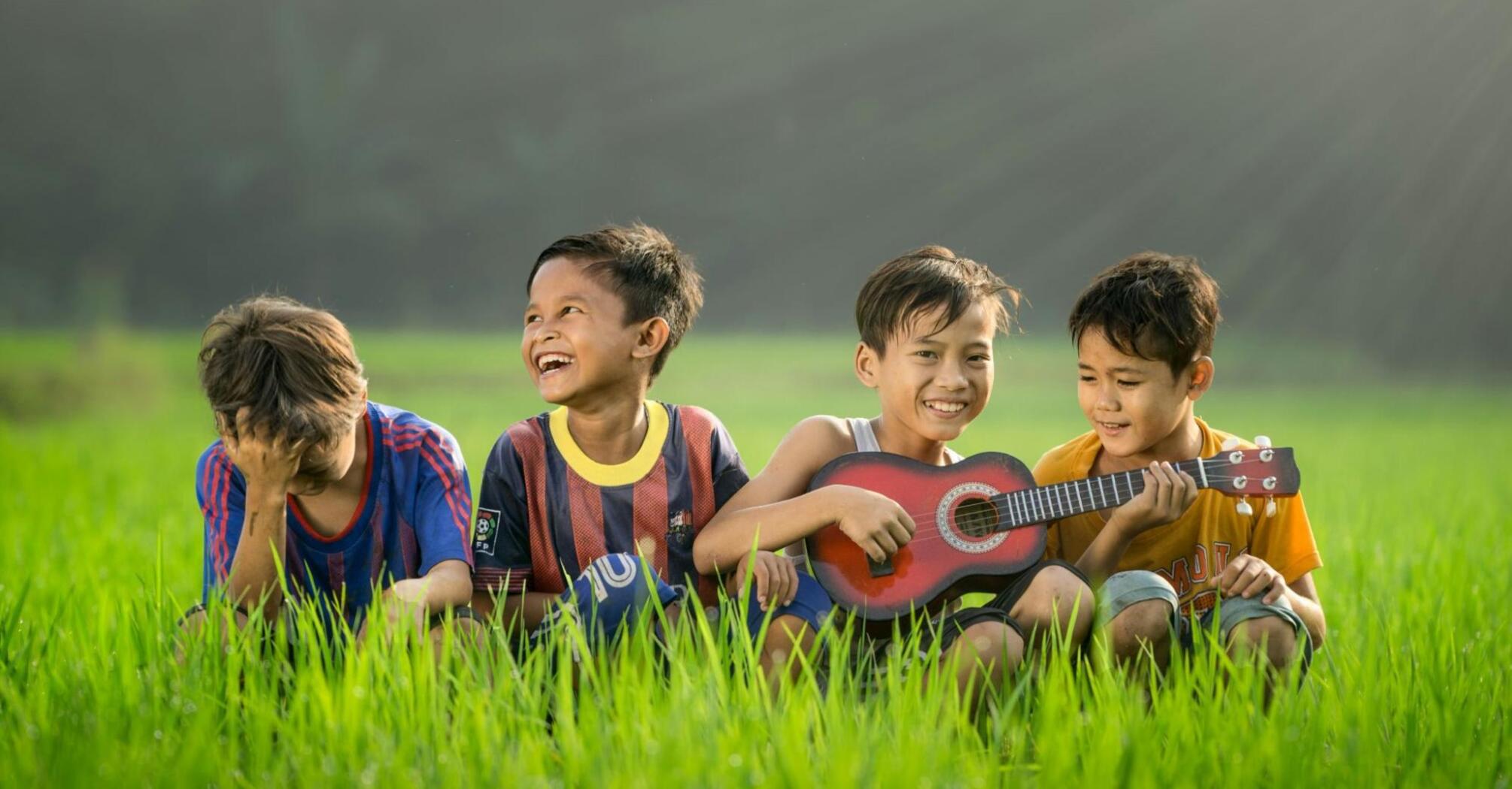 Four boys laugh on the grass, one plays the guitar