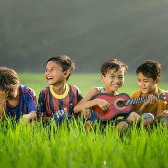 Four boys laugh on the grass, one plays the guitar