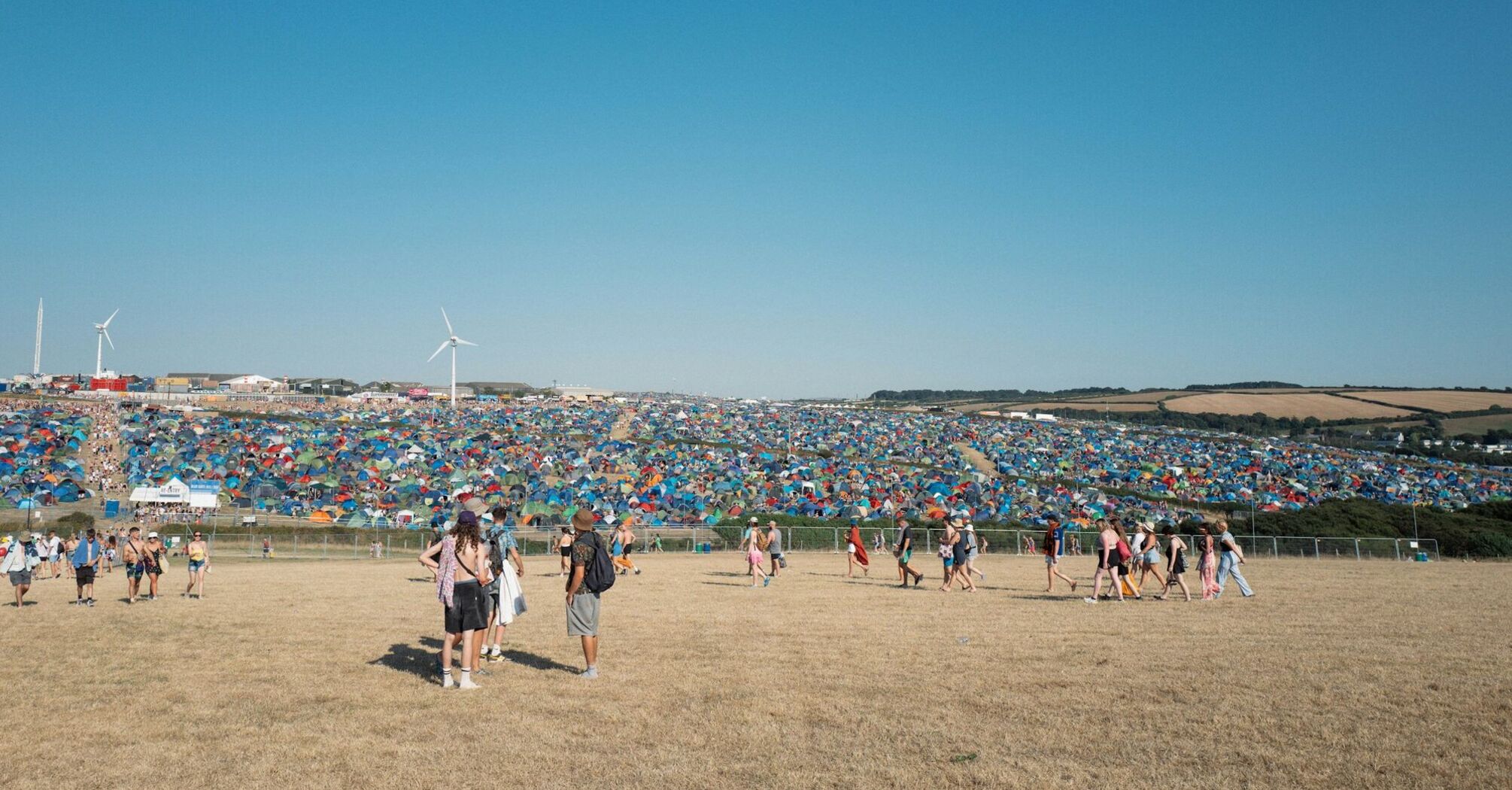 Festival goers walking towards a large campsite filled with colorful tents under a clear blue sky at Boardmasters