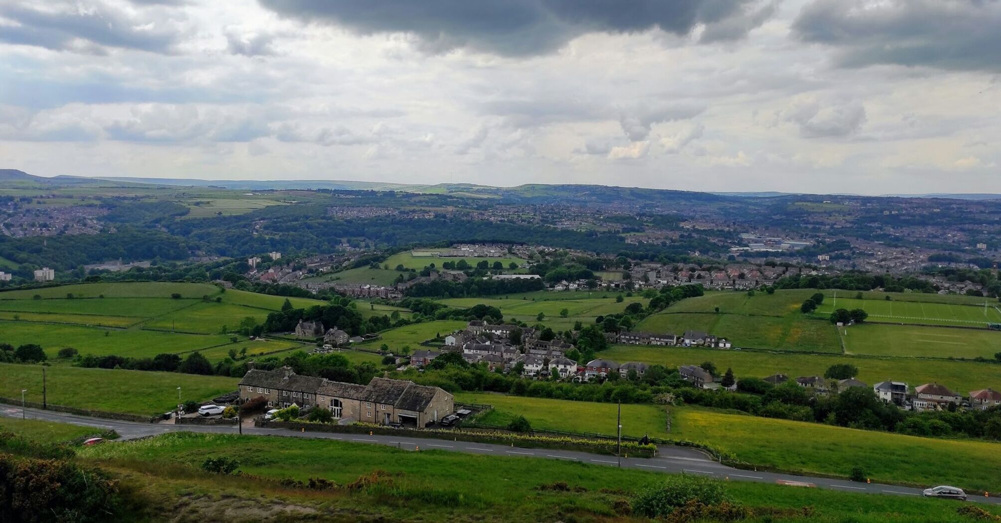 A scenic view of Huddersfield, showcasing lush green fields, rolling hills, and clusters of buildings under a cloudy sky