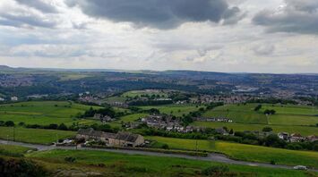 A scenic view of Huddersfield, showcasing lush green fields, rolling hills, and clusters of buildings under a cloudy sky