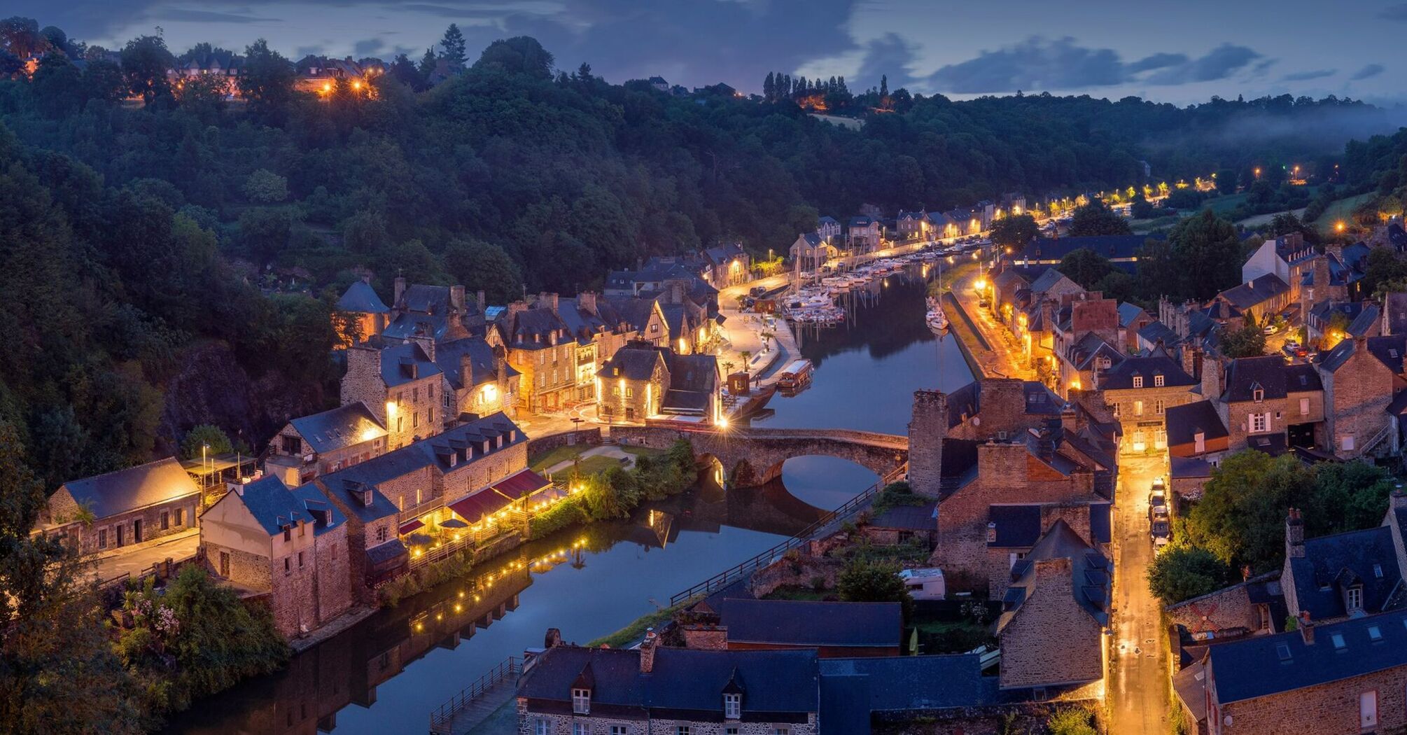 A picturesque evening view of a French village along a river, illuminated by street lights