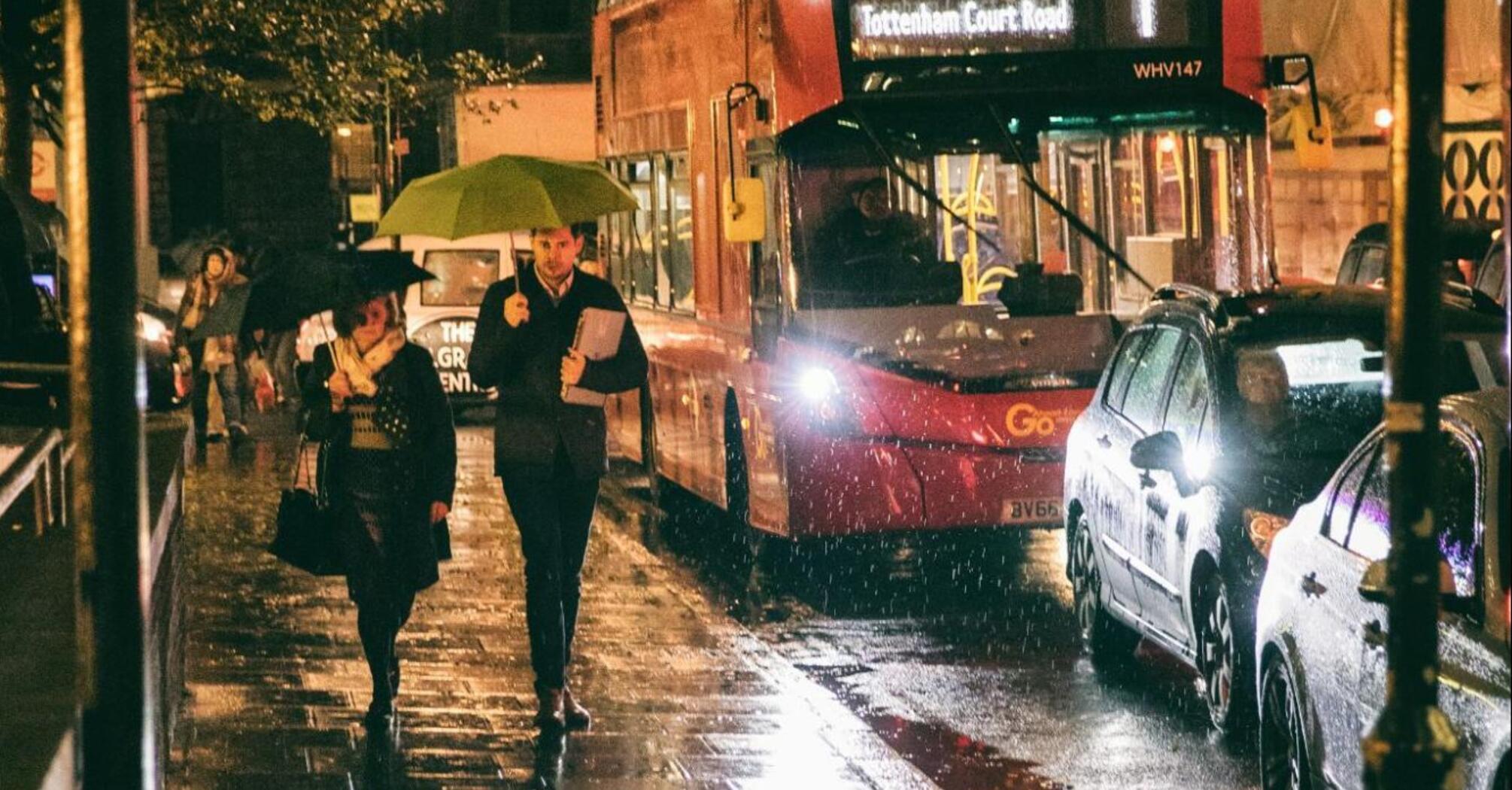 People walking with umbrellas on a rainy night near a bus stop with a red double-decker bus in the background
