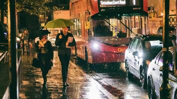 People walking with umbrellas on a rainy night near a bus stop with a red double-decker bus in the background