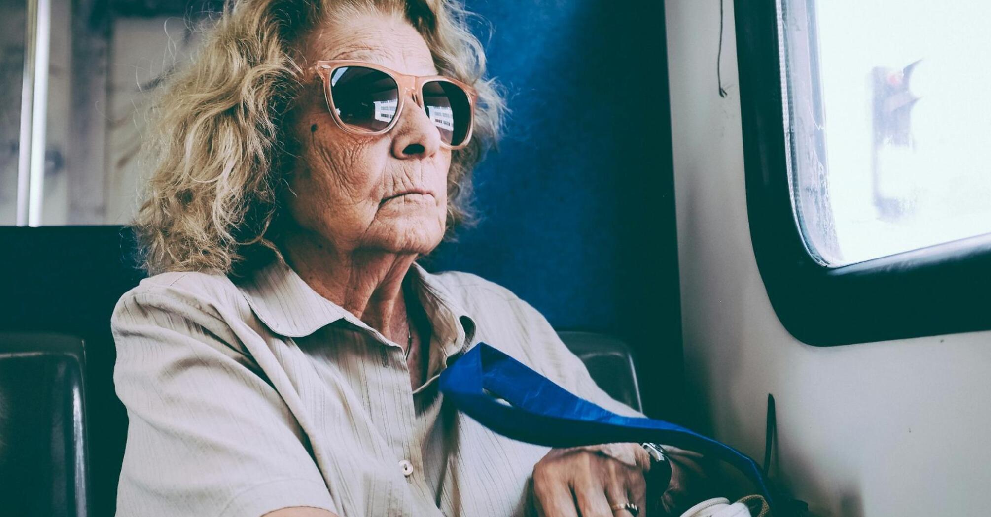An elderly woman wearing sunglasses sits on a bus, looking out the window