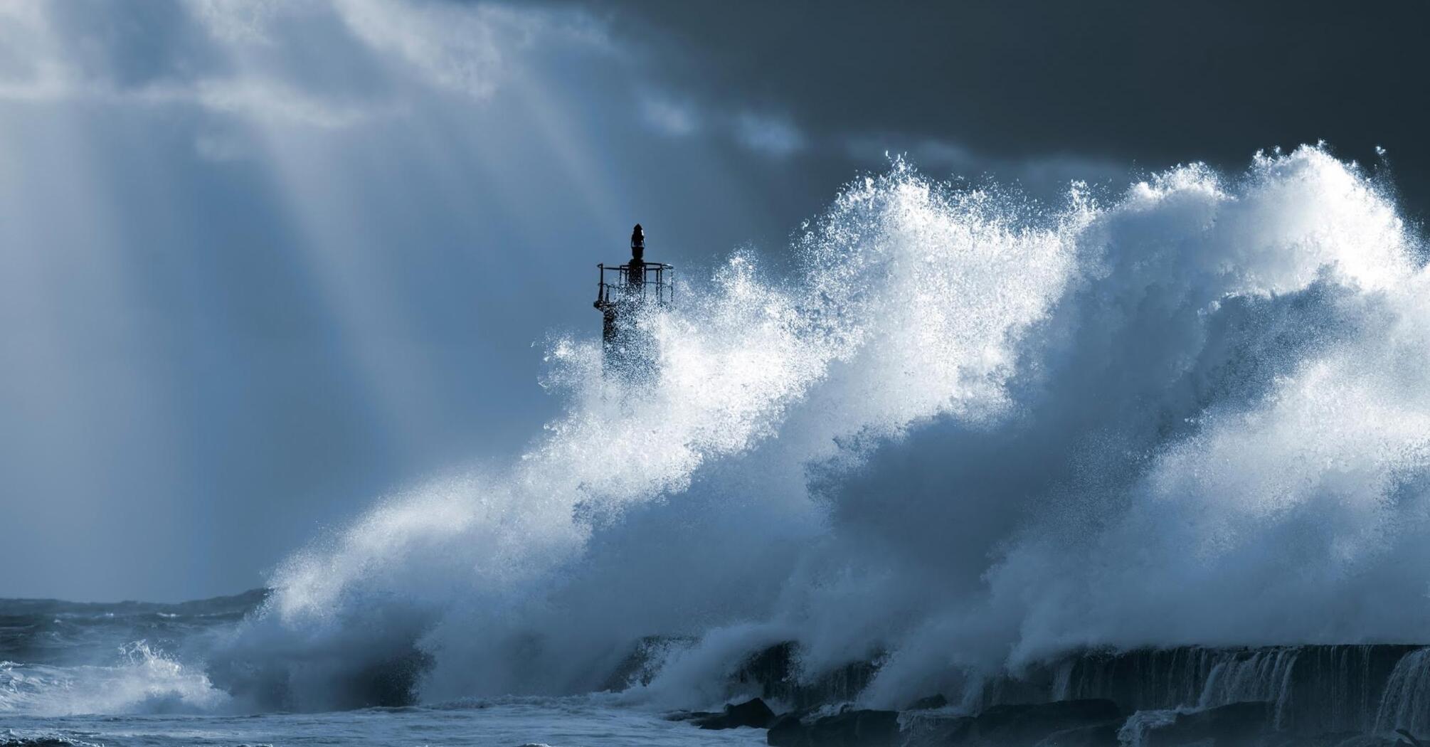 Powerful ocean waves crash against the lighthouse under a gloomy sky, creating a magnificent spray of water
