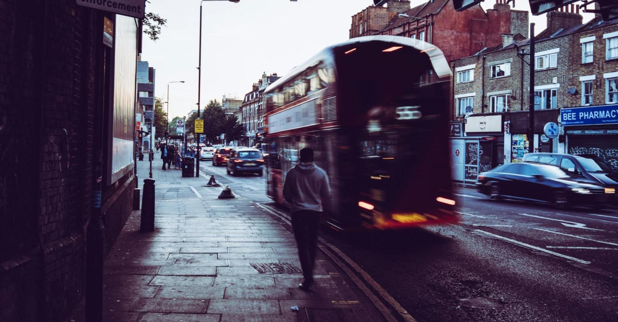 Blurred image of a red double-decker bus moving along a street with a pedestrian walking on the sidewalk