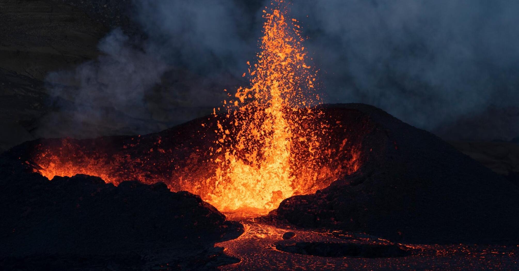 A powerful stream of lava bursts from the crater, sending a column of molten fragments into the sky