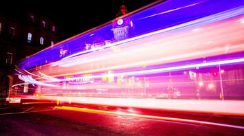 Long exposure photo of a night bus passing by a brightly lit building with a clock tower