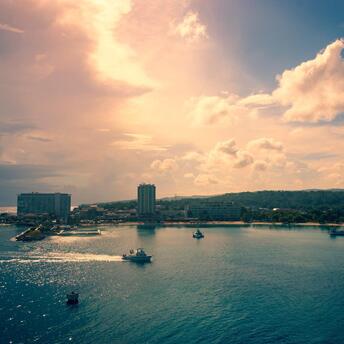 A tranquil view of boats navigating calm waters as the sun sets behind a coastal city, casting a warm glow over the scene