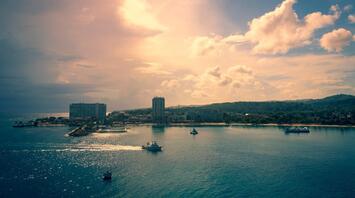 A tranquil view of boats navigating calm waters as the sun sets behind a coastal city, casting a warm glow over the scene