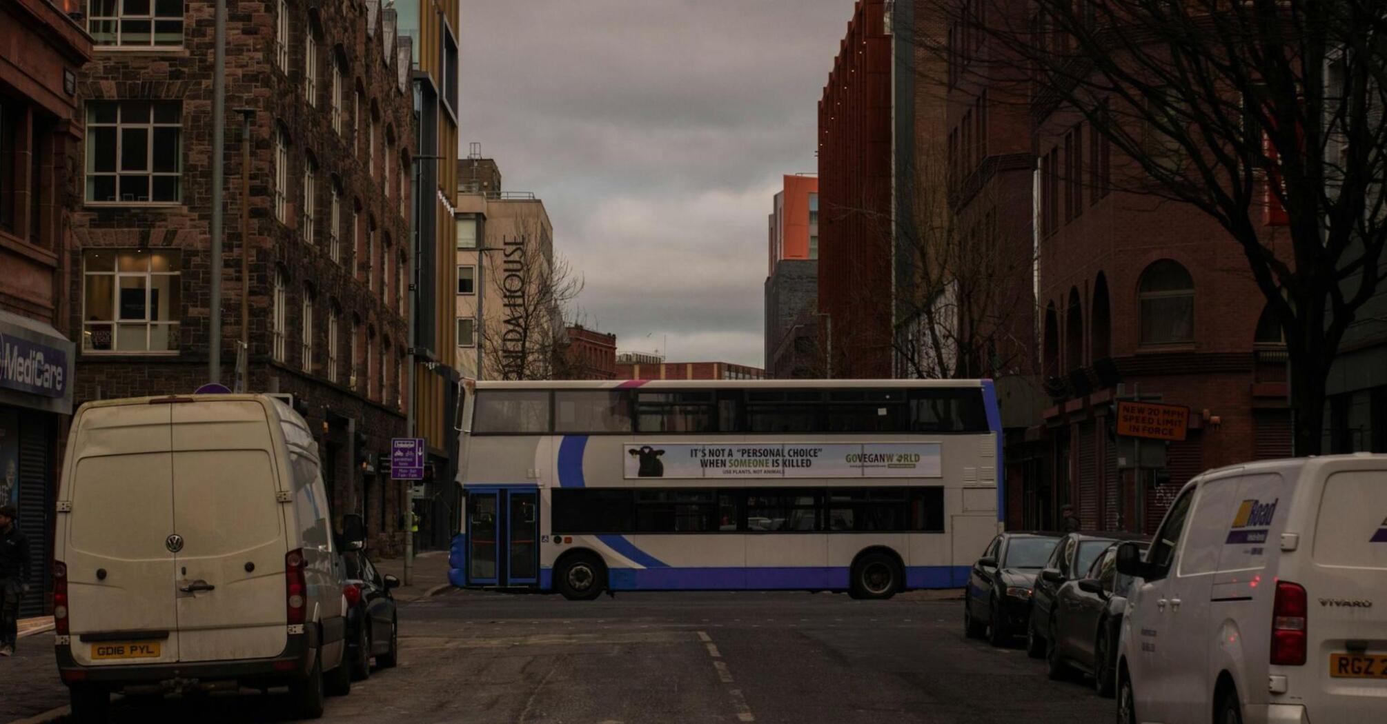 A city bus passes through a street lined with buildings, with a cloudy sky overhead
