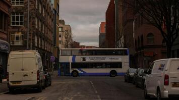 A city bus passes through a street lined with buildings, with a cloudy sky overhead