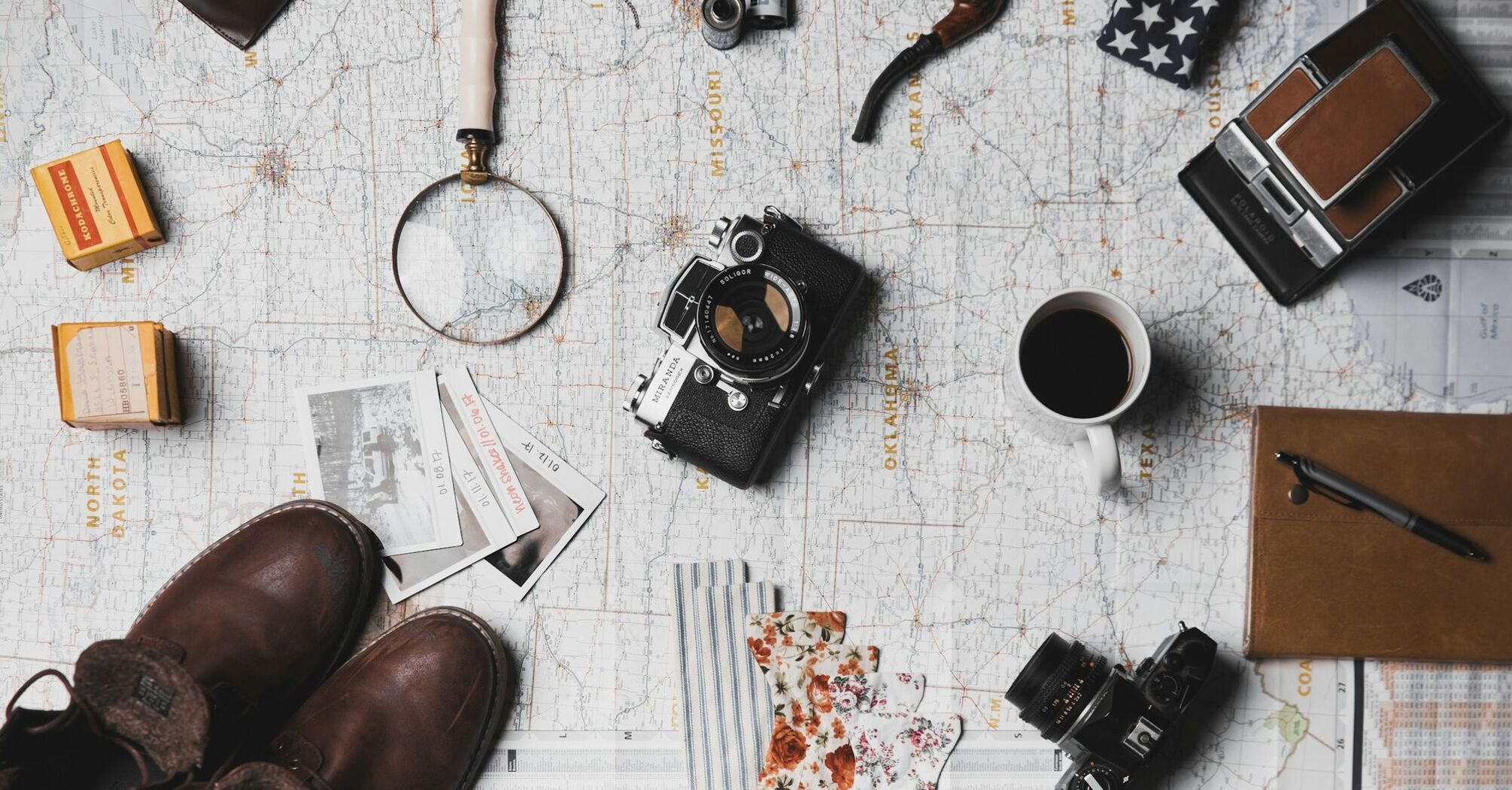 Travel essentials laid out on a map, including a camera, magnifying glass, boots, and a coffee cup