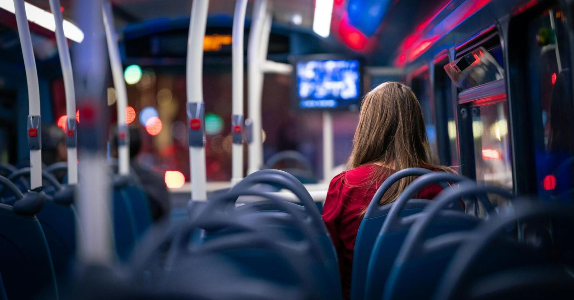 A woman sits alone on a nearly empty bus at night, with city lights visible through the windows