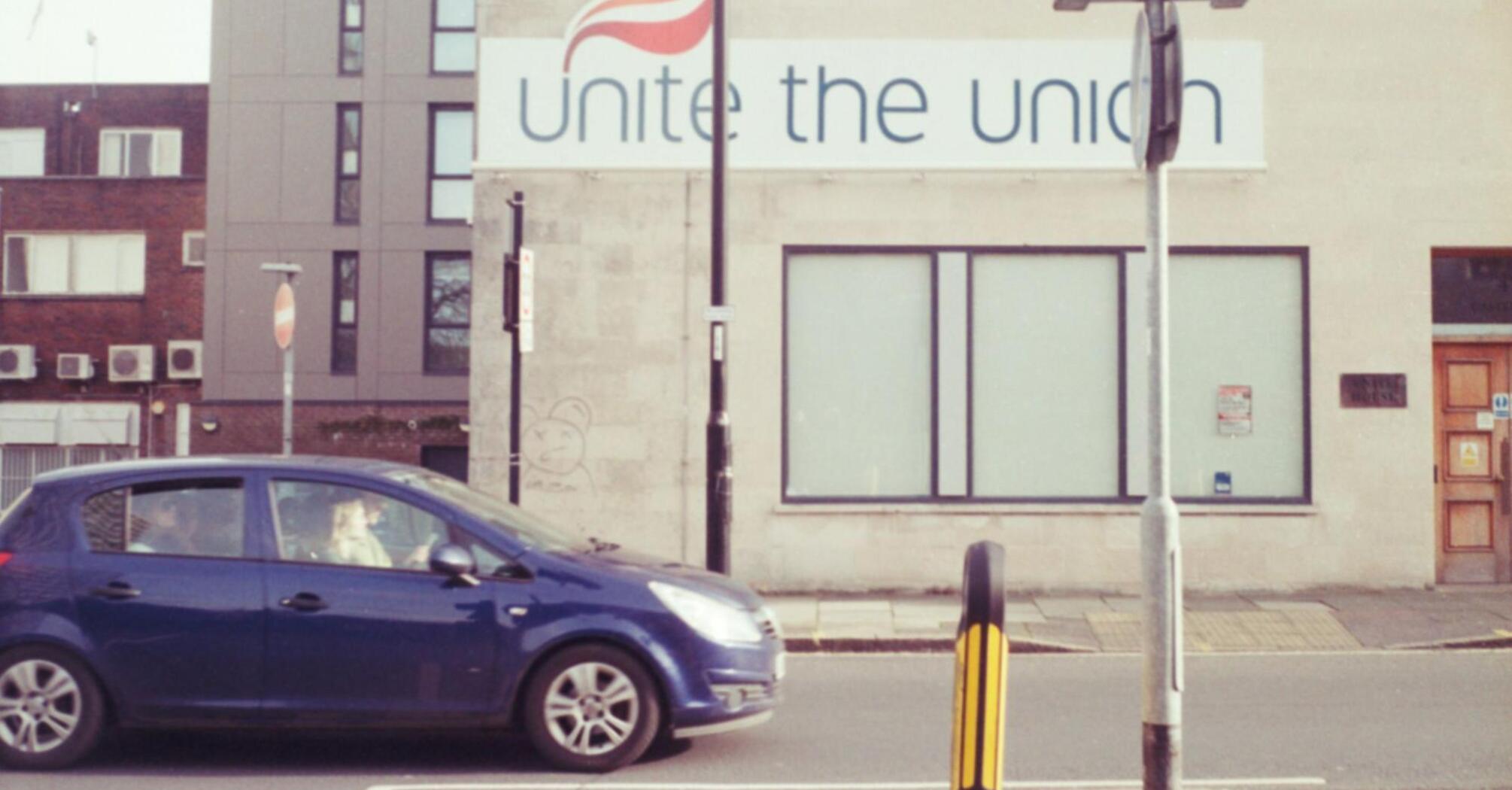 A blue car passing by a building with a large 'Unite the Union' sign