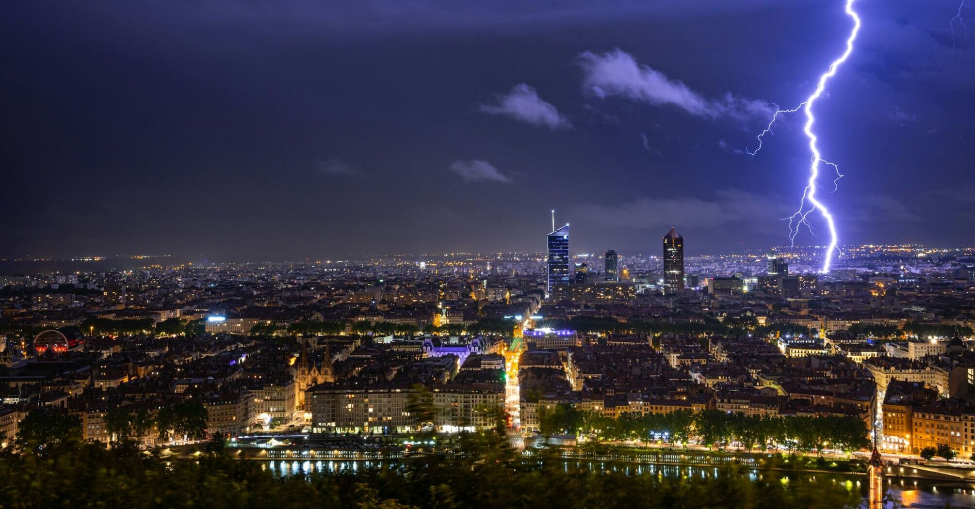 A dramatic lightning strike illuminates the night sky over Lyon, highlighting the skyline and buildings below