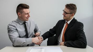 Two men in business attire shake hands while seated at a table, with a contract and pen in front of them