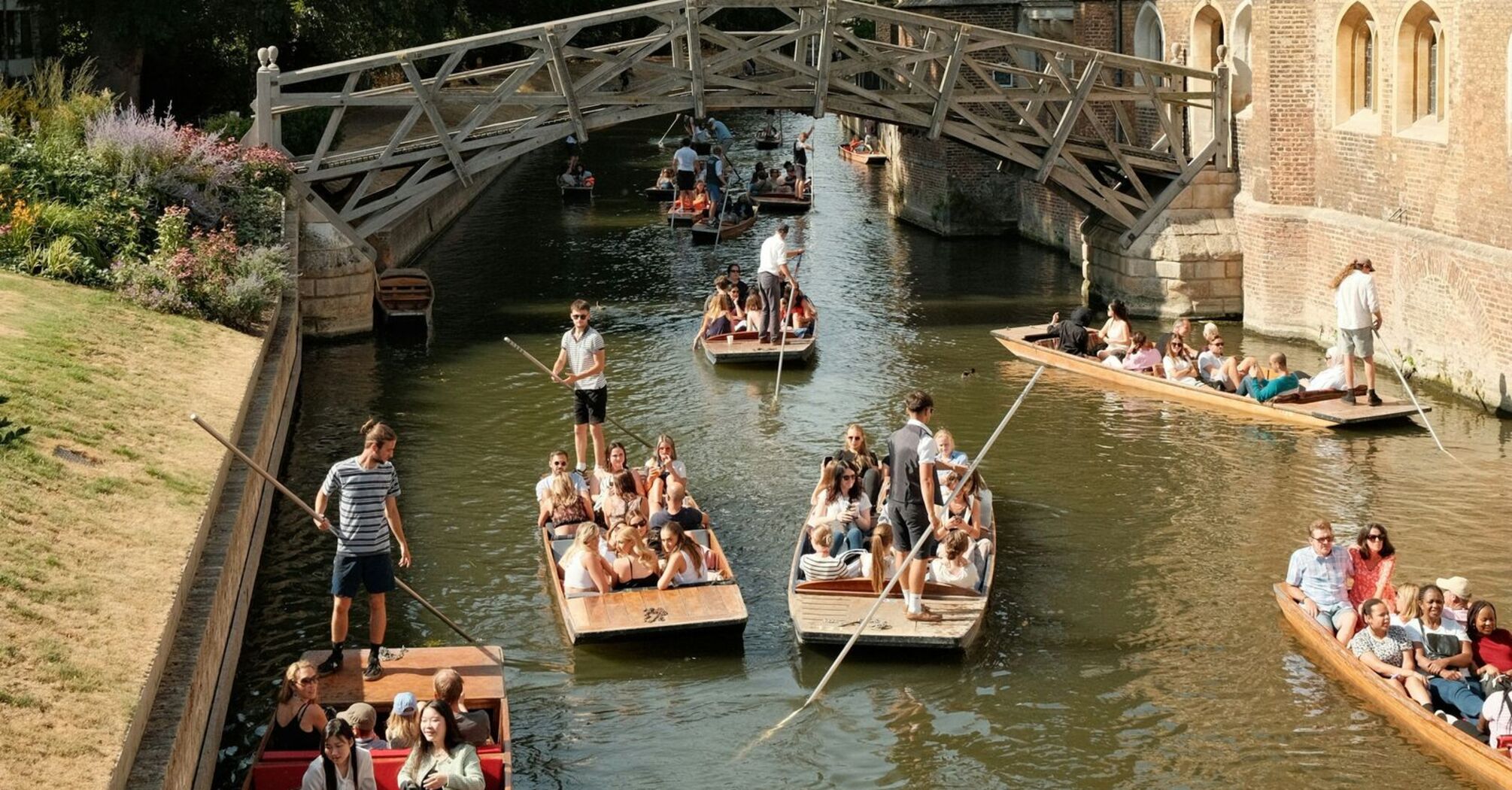 A group of tourists enjoy punting on the River Cam, passing under the historic Mathematical Bridge in Cambridge, surrounded by lush greenery and historic college buildings