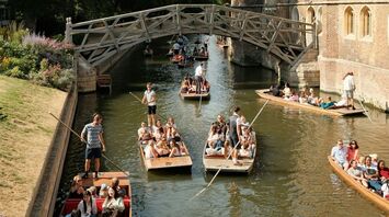 A group of tourists enjoy punting on the River Cam, passing under the historic Mathematical Bridge in Cambridge, surrounded by lush greenery and historic college buildings
