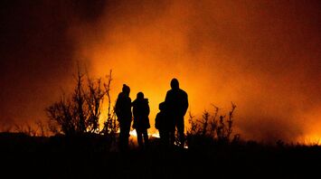 Silhouetted figures standing near intense wildfire at night