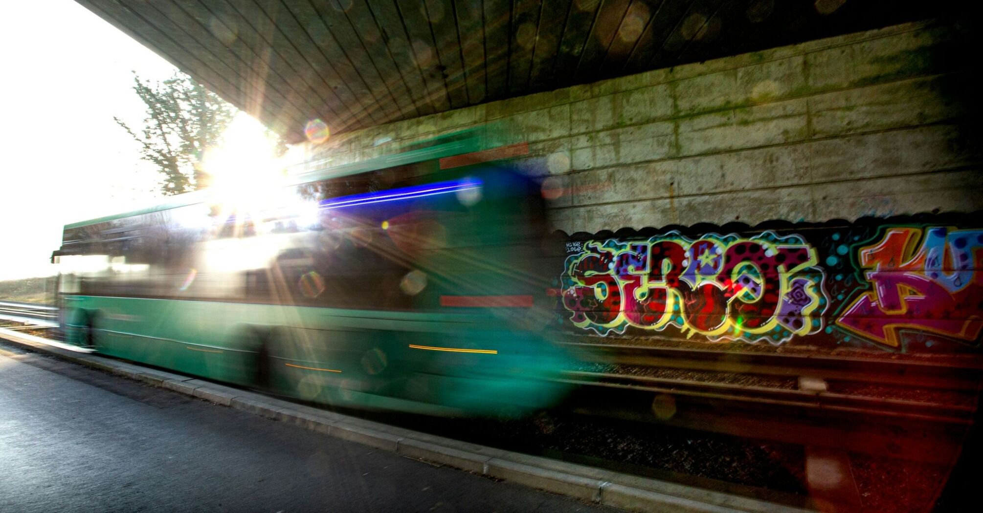 A green bus speeds under a graffiti-covered bridge with sunlight flaring in the background