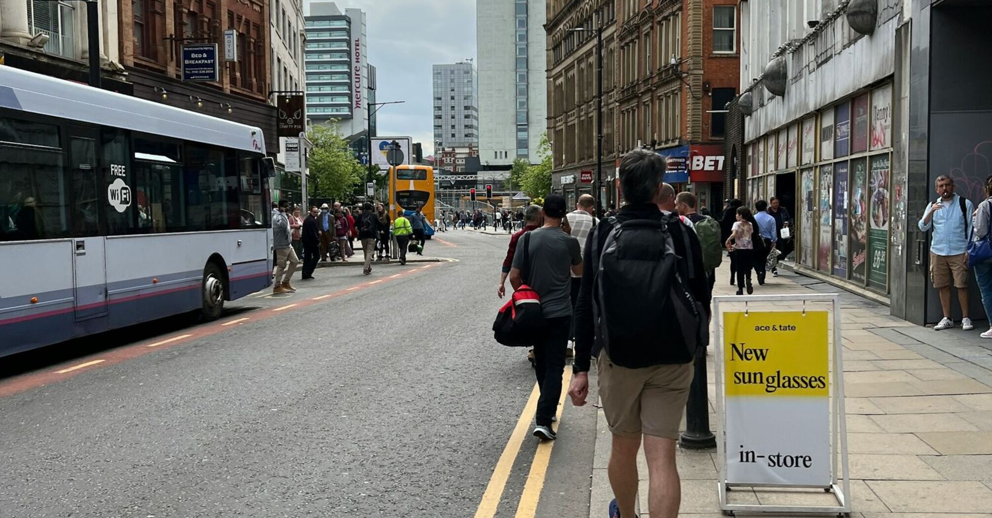 A busy street in Manchester with pedestrians walking alongside buildings and buses driving on the road