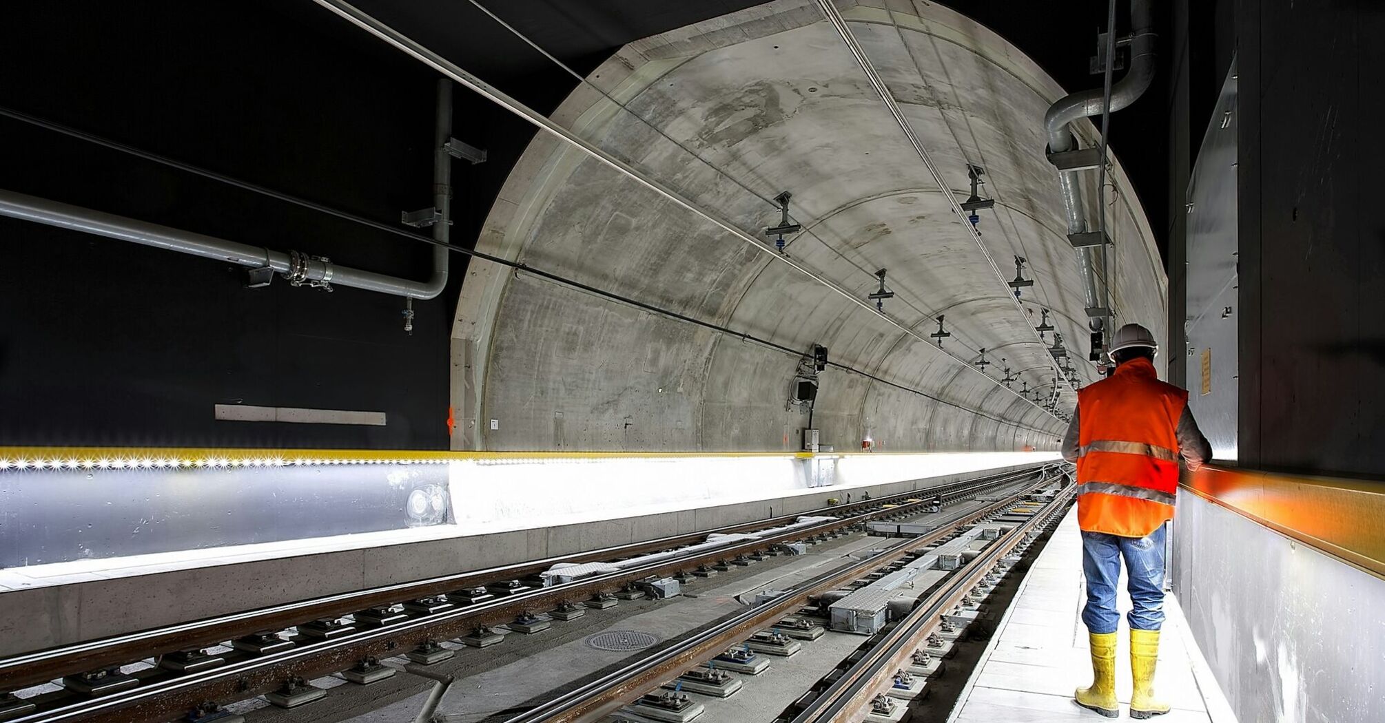 A worker in a reflective vest and yellow boots stands inside a railway tunnel, inspecting the tracks and infrastructure