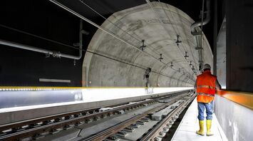 A worker in a reflective vest and yellow boots stands inside a railway tunnel, inspecting the tracks and infrastructure