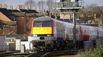 A Transport for Wales train approaches a station on a sunny day, with residential buildings in the background
