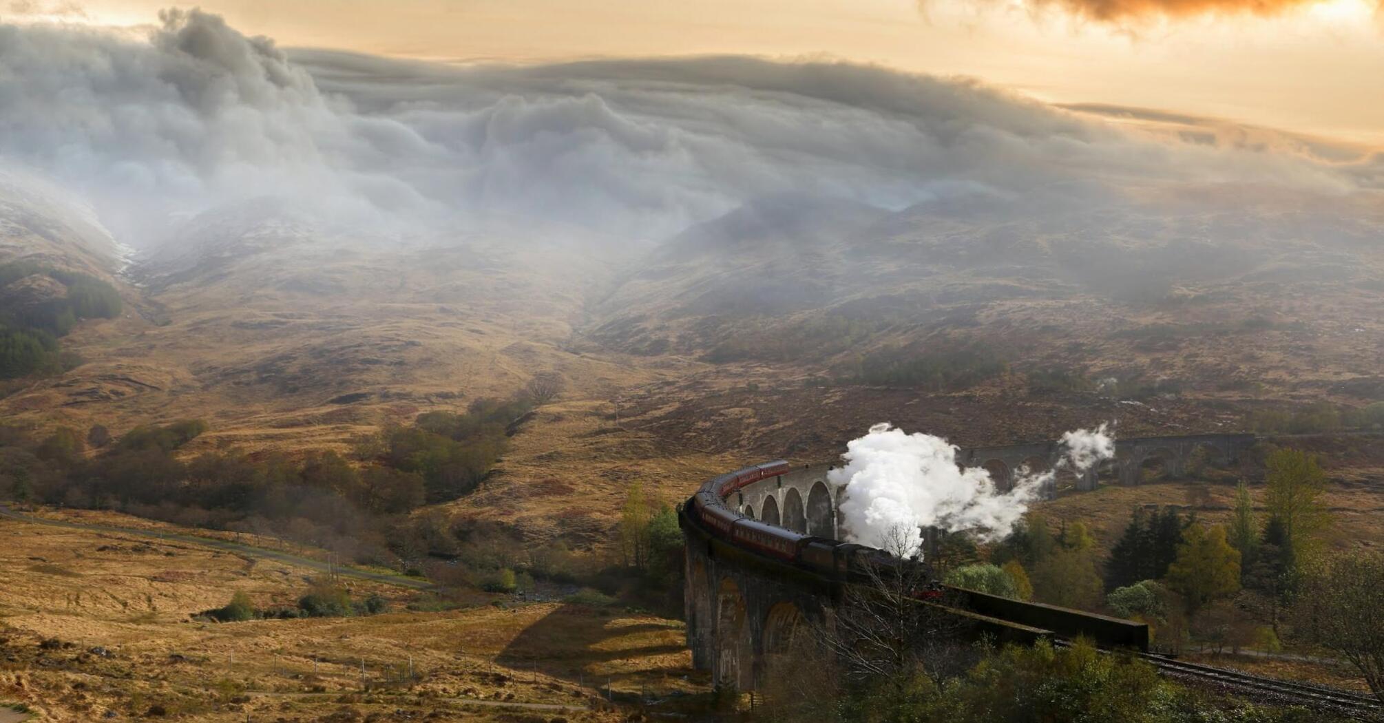 A steam train crosses a viaduct in a misty mountain landscape at dawn