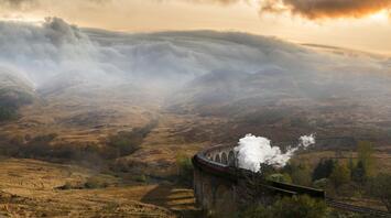 A steam train crosses a viaduct in a misty mountain landscape at dawn