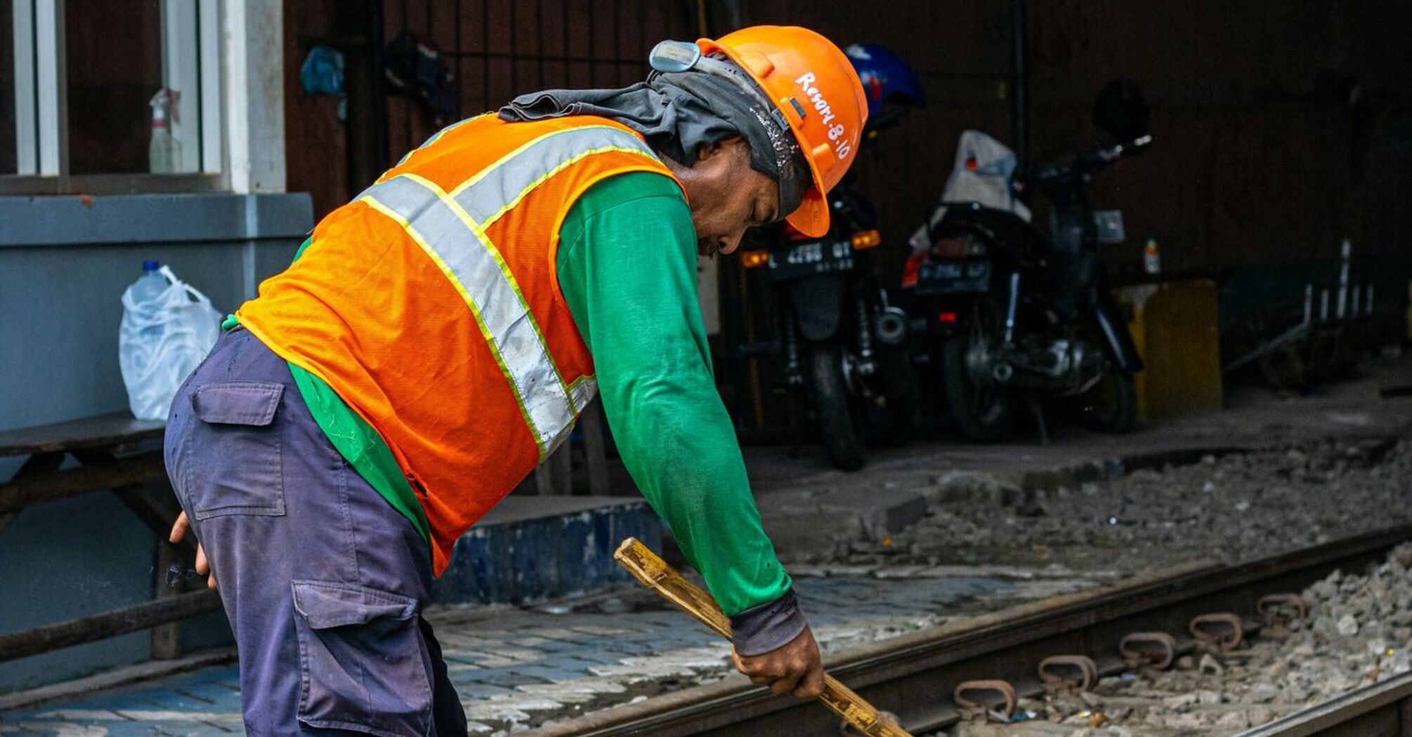 A railway worker in a high-visibility vest and helmet uses a hammer to work on railway tracks during maintenance