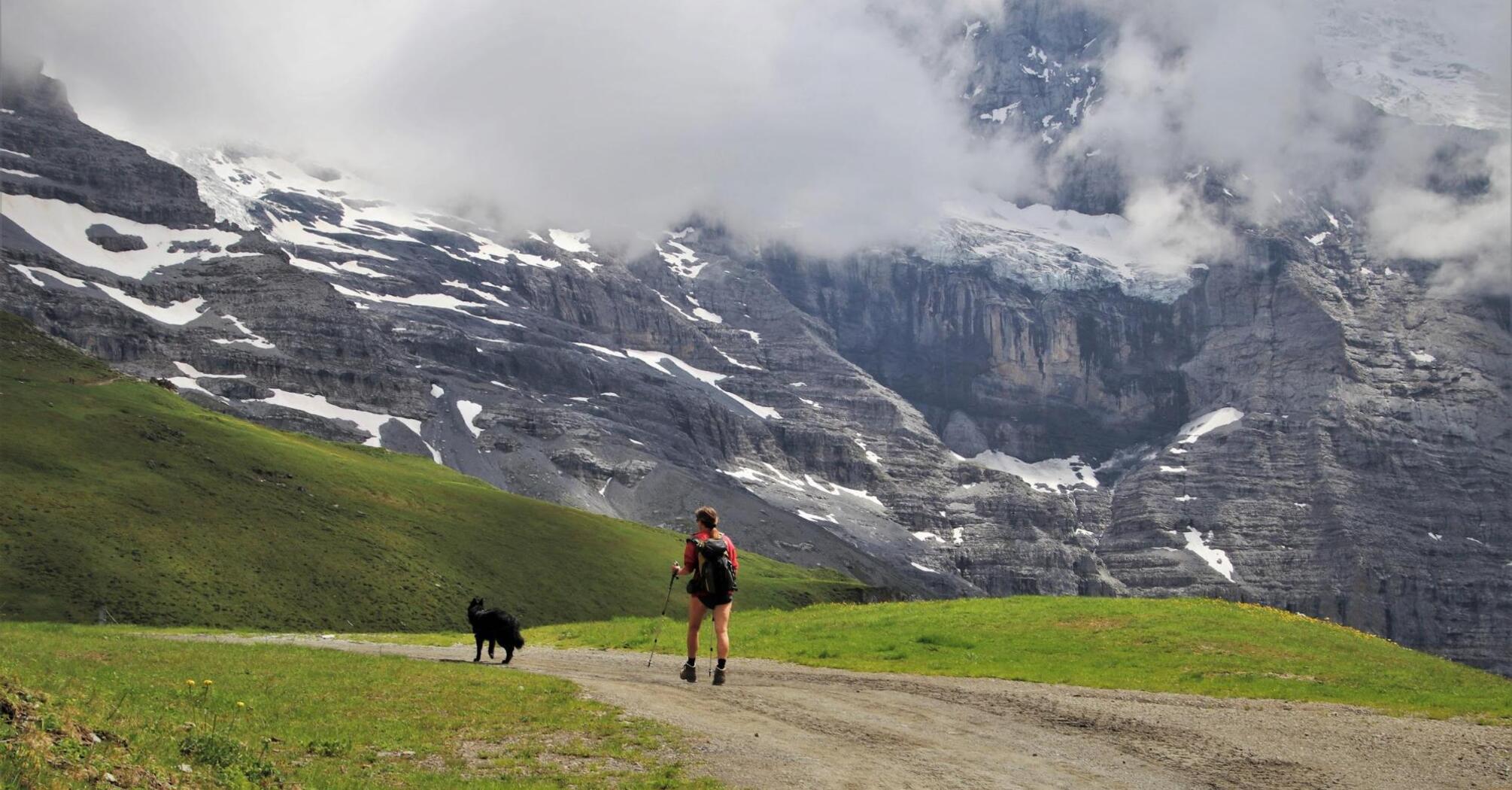 A hiker with a dog walking along a mountain trail with snow-capped peaks and clouds in the background