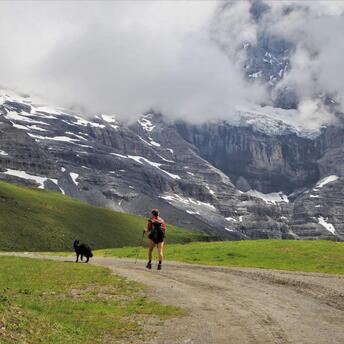 A hiker with a dog walking along a mountain trail with snow-capped peaks and clouds in the background