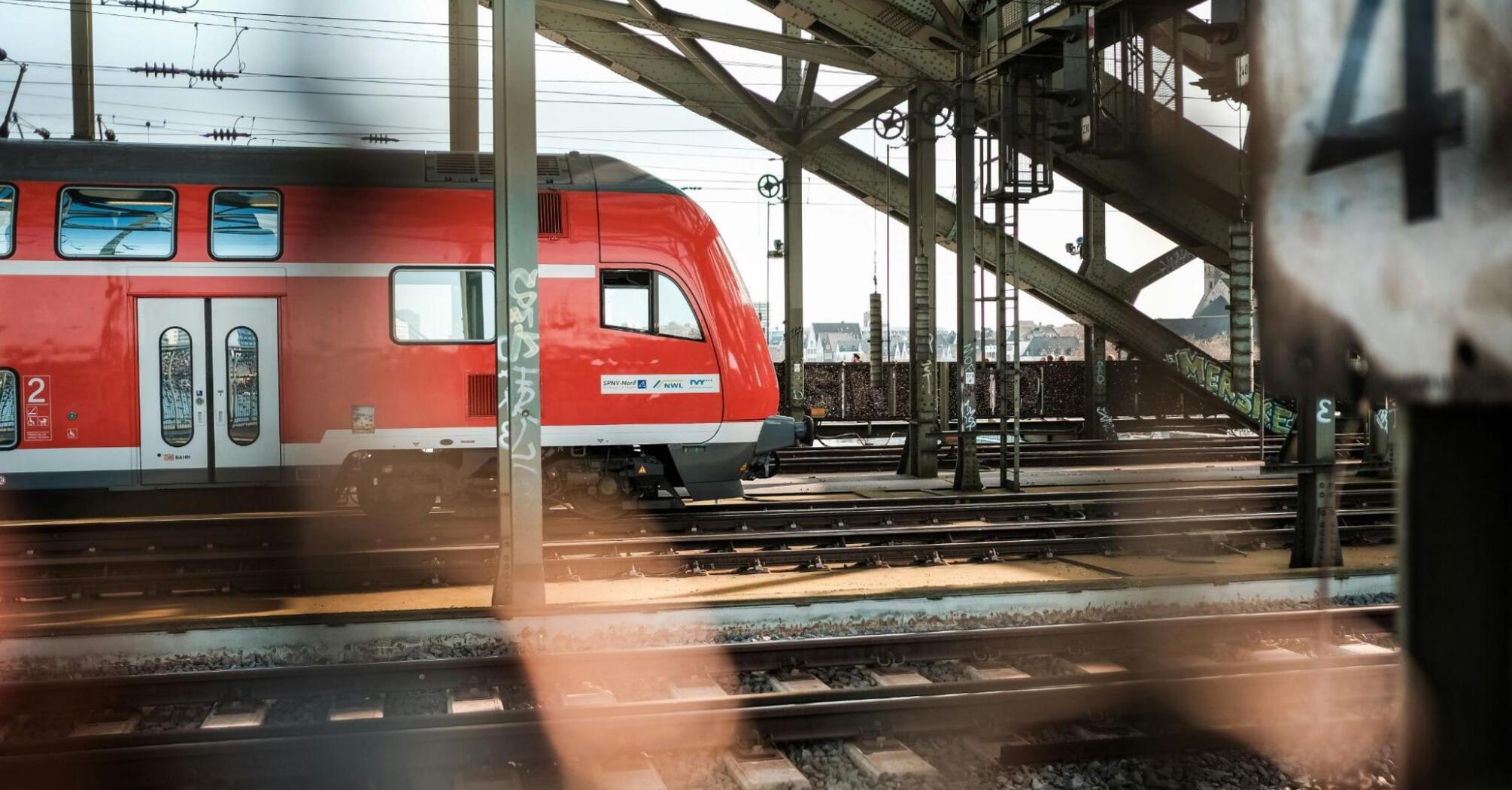 A red regional train stands idle on the tracks under a steel railway bridge, highlighting the temporary disruptions on the Berlin-Hamburg route