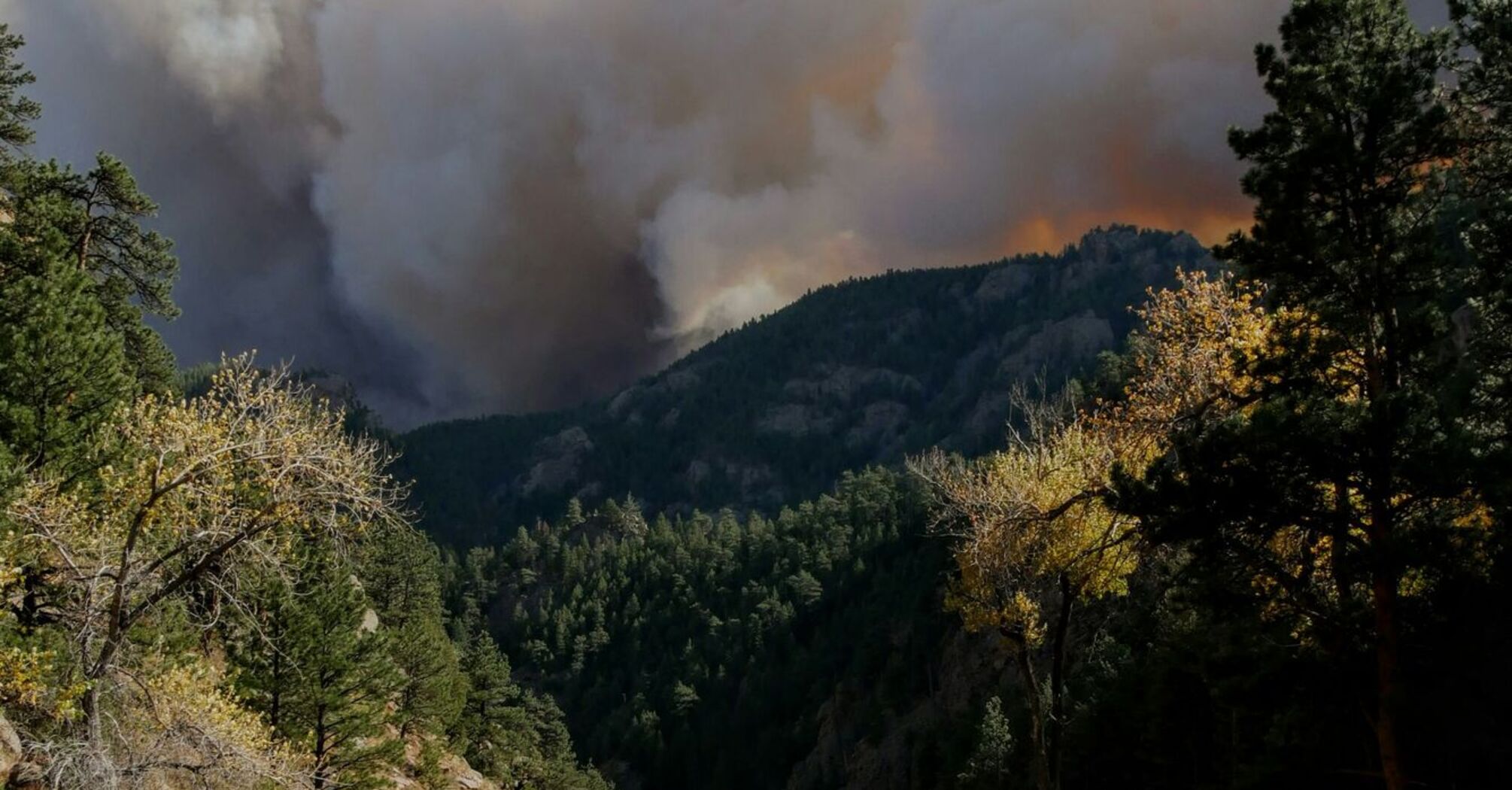 A large plume of smoke rises above a forested mountain area as a wildfire burns in the distance, with a highway running through the scene