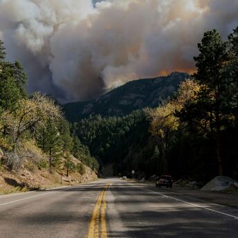 A large plume of smoke rises above a forested mountain area as a wildfire burns in the distance, with a highway running through the scene