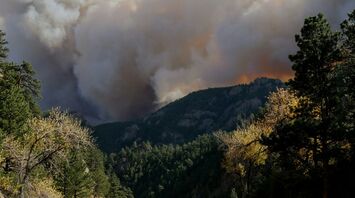 A large plume of smoke rises above a forested mountain area as a wildfire burns in the distance, with a highway running through the scene
