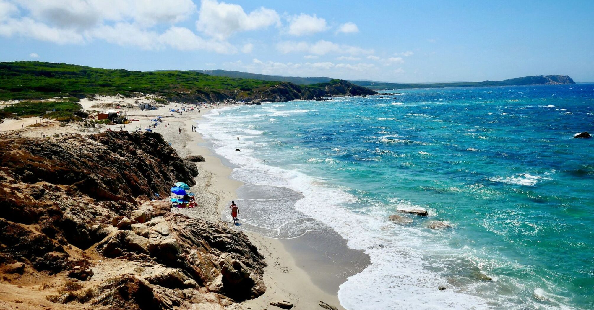 A Sardinia beach scene with turquoise waters, rocky cliffs, and people enjoying the sandy shoreline under a bright, clear sky