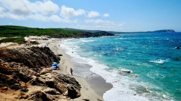 A Sardinia beach scene with turquoise waters, rocky cliffs, and people enjoying the sandy shoreline under a bright, clear sky
