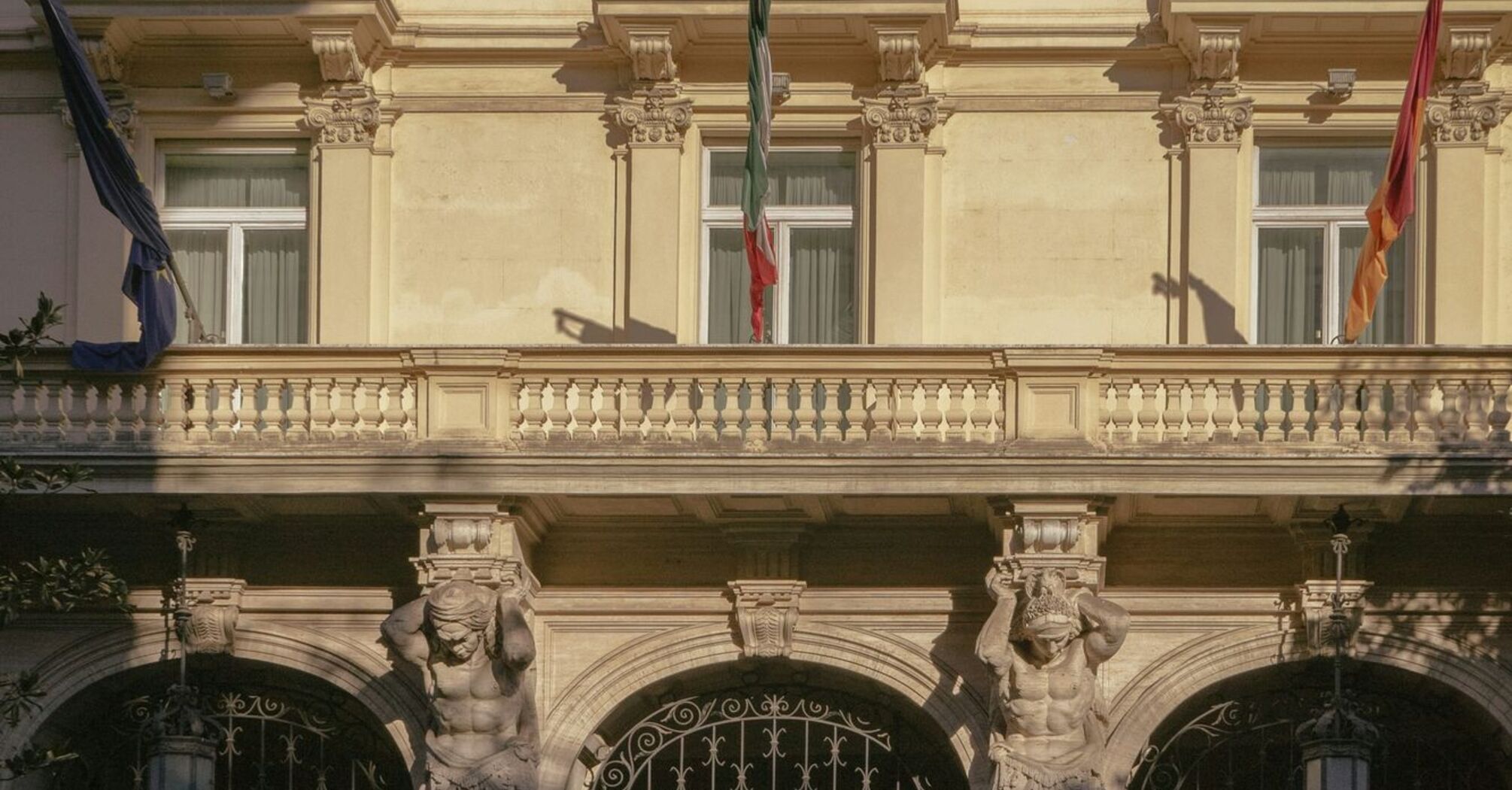 The facade of a historic luxury hotel in Rome with ornate architectural details and flags displayed above its entrance