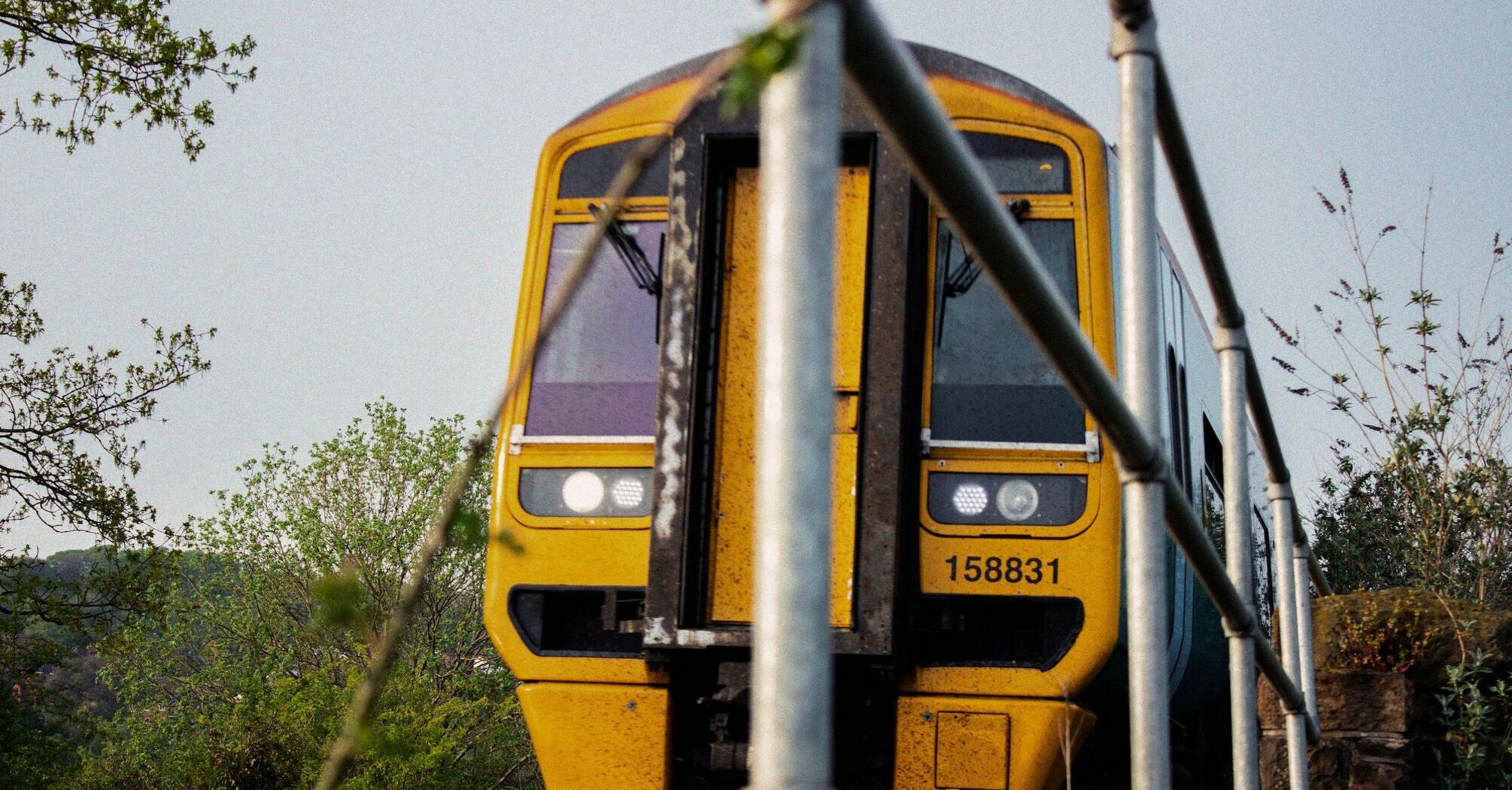 A yellow train captured from the front, partially obscured by metal railings, traveling through a rural area with greenery in the background