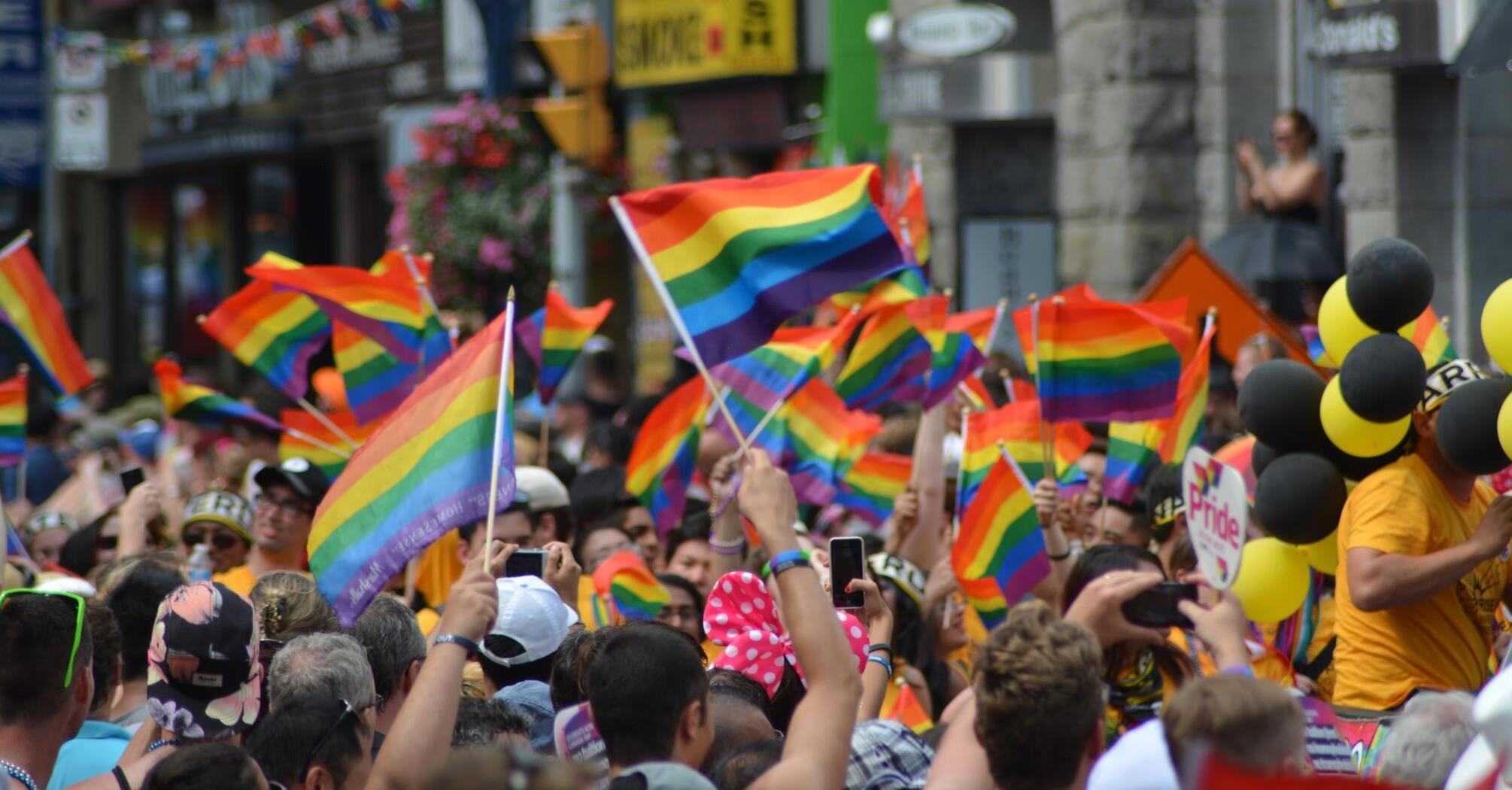 A crowded street filled with people waving rainbow flags during a Pride parade, with colorful storefronts and balloons in the background.