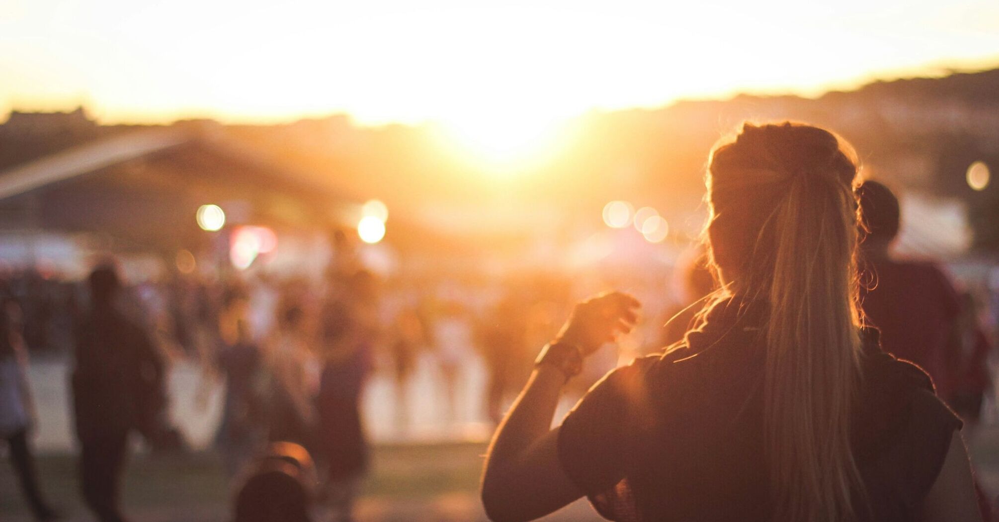 A crowd enjoying a festival at sunset, with a woman in the foreground facing away, capturing the vibrant and warm atmosphere